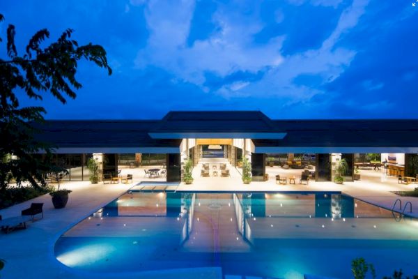 The image shows a luxurious, illuminated building with a large outdoor pool in the foreground, set against a twilight sky.