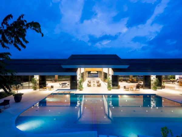 The image shows a luxurious, illuminated building with a large outdoor pool in the foreground, set against a twilight sky.