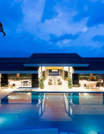 A luxurious, illuminated building with a large outdoor pool reflects the evening sky.