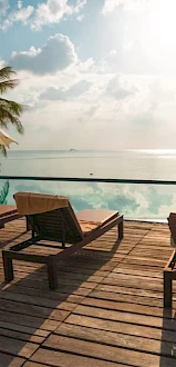 A serene poolside scene with lounge chairs, umbrellas, palm trees, and a view of the ocean under a cloudy sky.