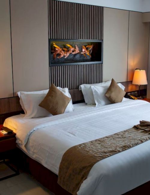 A cozy hotel room with a large bed, two lamps, decorative pillows, and a wall-mounted fireplace, creating a warm ambiance.