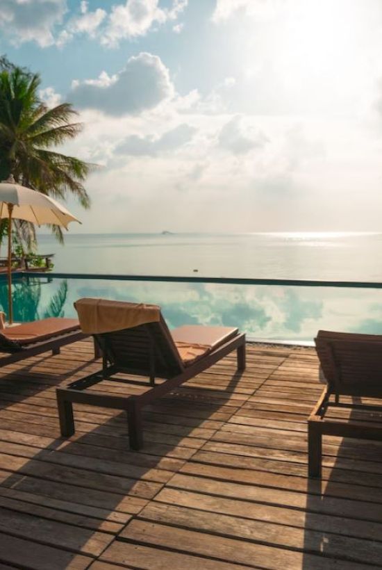 A serene poolside scene with lounge chairs, umbrellas, palm trees, and a view of the ocean under a partially cloudy sky.