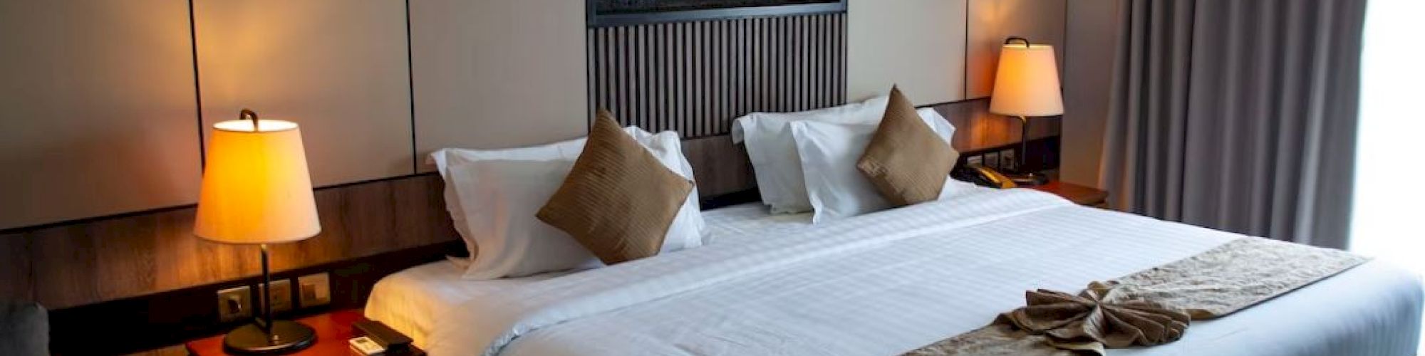 A cozy hotel room features a large bed with decorative pillows, warm lighting, bedside tables, and a modern wall-mounted fireplace.