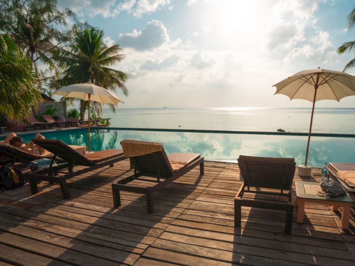 A serene poolside with lounge chairs, umbrellas, and palm trees overlooking a calm, sunlit sea under a partly cloudy sky.