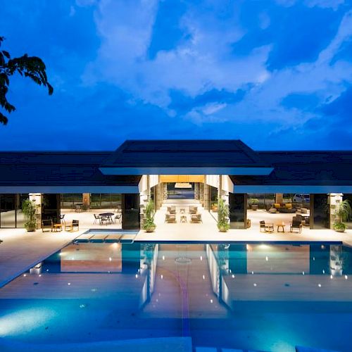 A luxurious, illuminated pool area at night with a large modern building in the background under a partly cloudy sky.