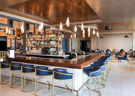 A modern bar with wood accents, stylish lighting, blue barstools, and a well-stocked back bar. Lounge seating is visible in the background.