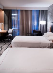 A modern hotel room with two beds, a TV, desk, chair, and ambient lighting. The decor is sleek, featuring neutral tones and patterns.