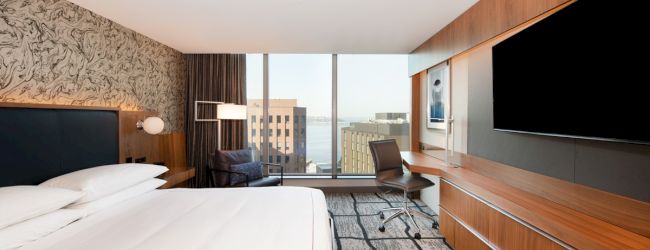 A modern hotel room with a large bed, desk, chair, and TV, featuring a window view of buildings and water in the background.