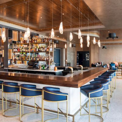 A stylish bar with blue stools, a stocked liquor shelf, and hanging lights, in a modern lounge setting with seating areas.