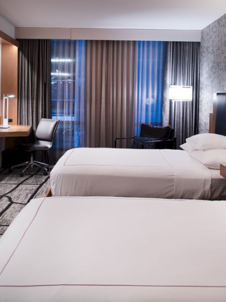 A modern hotel room with two beds, a wall-mounted TV, desk, chair, and large window with curtains.