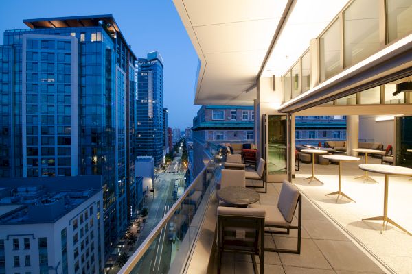 A modern rooftop balcony overlooking an urban street at dusk, with tables and chairs, and tall buildings in the background.