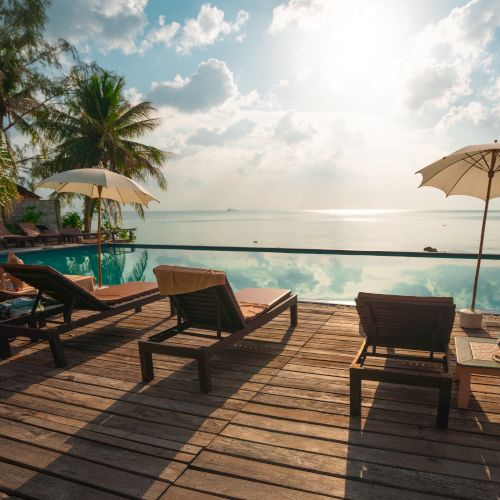 A serene beach scene with lounge chairs, umbrellas, and a pool overlooking the ocean, under a bright sun and partly cloudy sky.