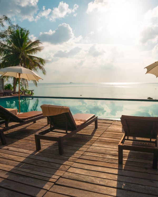 A serene beach scene with lounge chairs, umbrellas, and a pool overlooking the ocean, under a bright sun and partly cloudy sky.