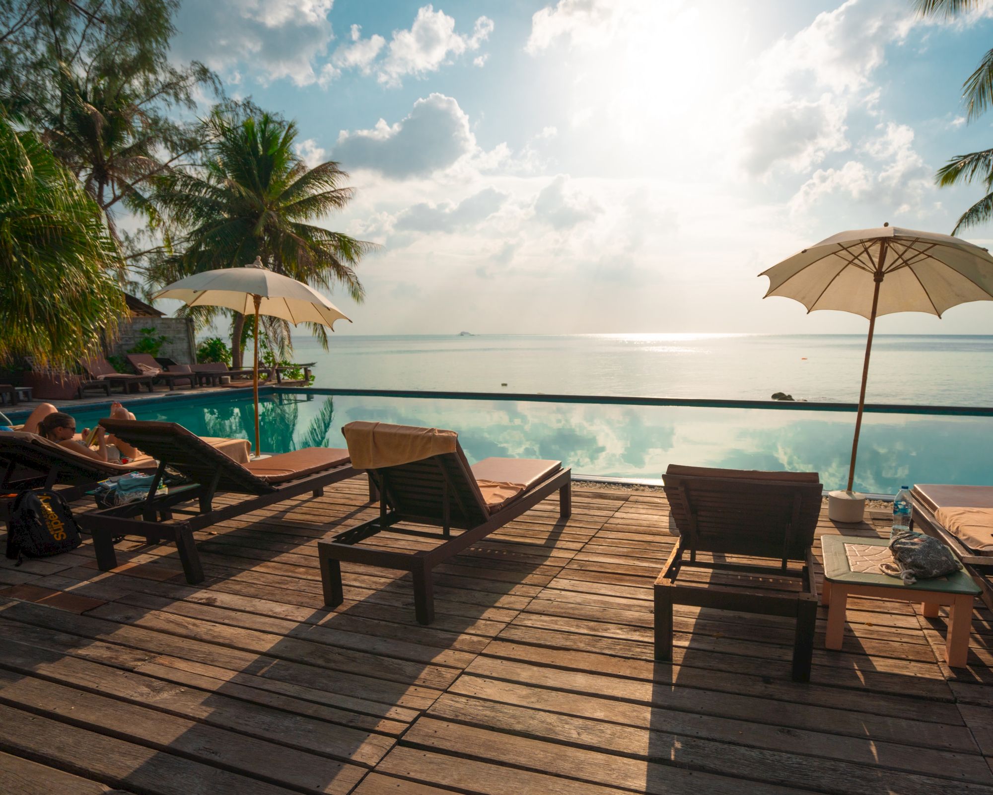 A serene poolside view with sun loungers, umbrellas, palm trees, and a tranquil sea under a bright sky and soft clouds.