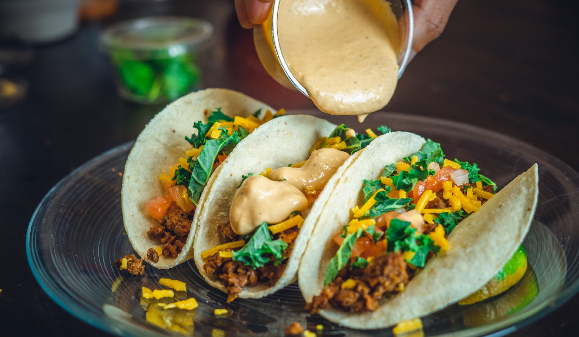 Three tacos filled with meat, cheese, tomatoes, and greens are being topped with creamy sauce from a cup.