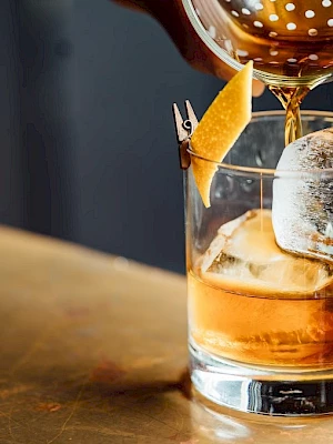 A cocktail being poured into a glass with ice and garnished with a twist of lemon peel.