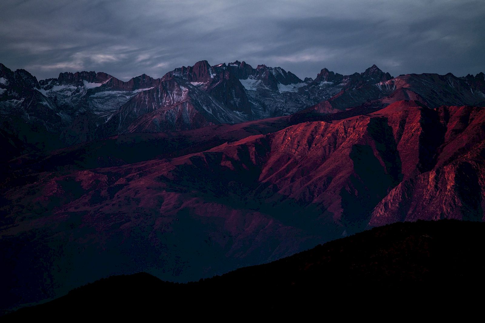 A rugged mountain range with snow-capped peaks is bathed in warm, red light under a dramatic sky.