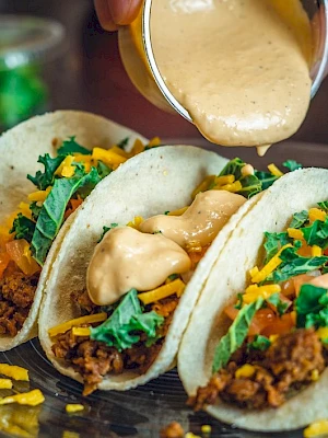 Three tacos filled with ground meat, cheese, lettuce, and tomatoes on a plate, with a sauce being poured over them.