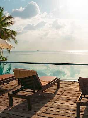 The image shows a tranquil seaside pool area with lounge chairs, umbrellas, and palm trees, overlooking a serene ocean view.