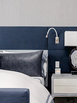A modern bedroom features a bed with blue and gray pillows, a nightstand, a lamp, and a wall-mounted reading light.