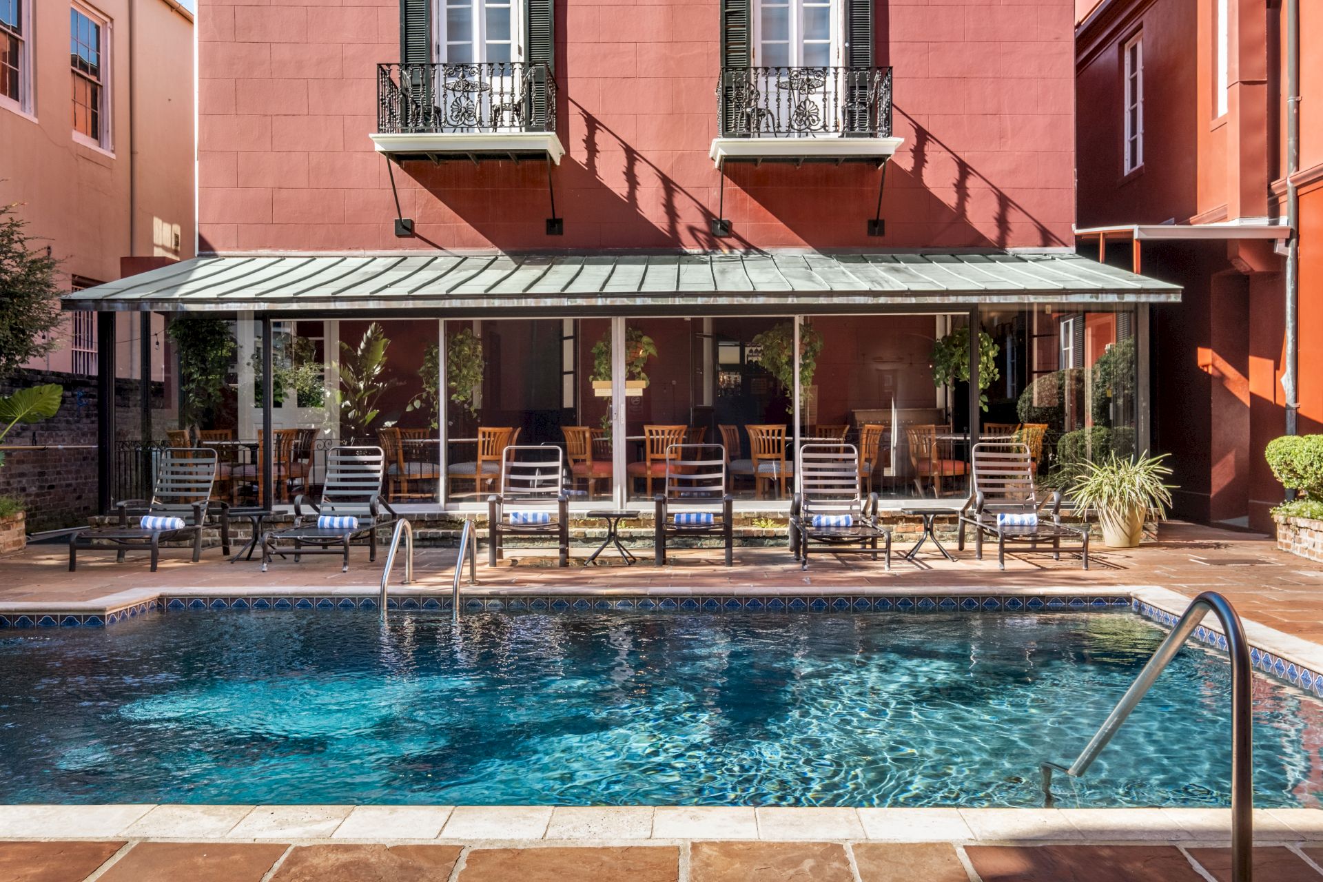 A swimming pool with lounge chairs, a glass-covered patio, and plants in front of a red building with balconies.