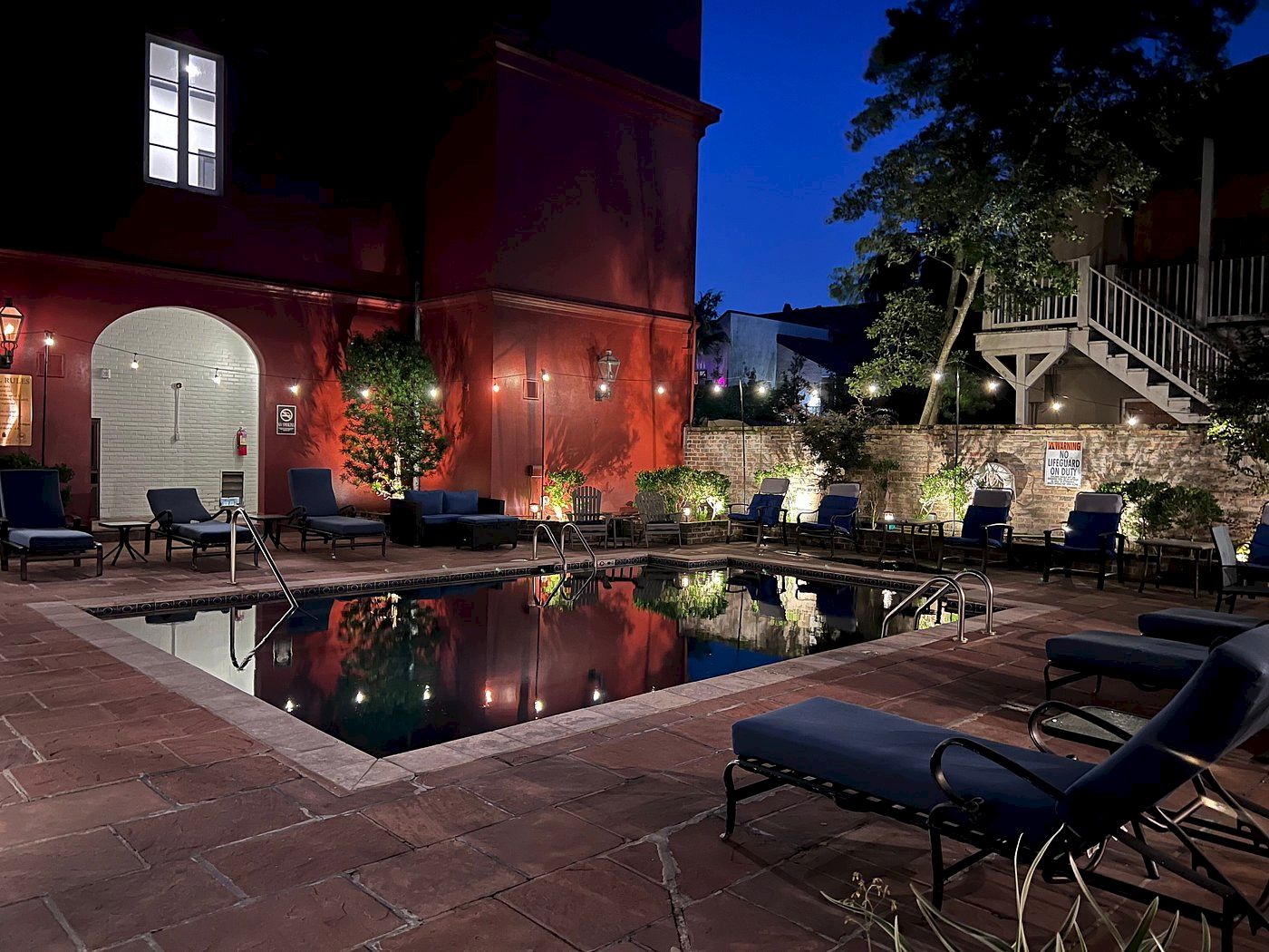 A nighttime scene of a tranquil outdoor courtyard features a pool surrounded by lounge chairs and a building with soft lighting.