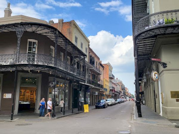 A street scene with historic buildings, featuring ironwork balconies, people walking, and cars parked along the road under a partly cloudy sky.
