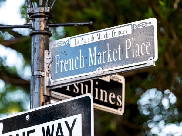 Street signs reading "French Market Place" and "Ursulines" with a "One Way" sign beneath, set against a leafy background.