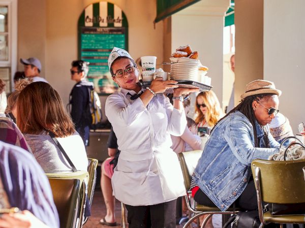 A waiter carries a tray through a busy outdoor cafe, with people seated at tables enjoying their meals and drinks.