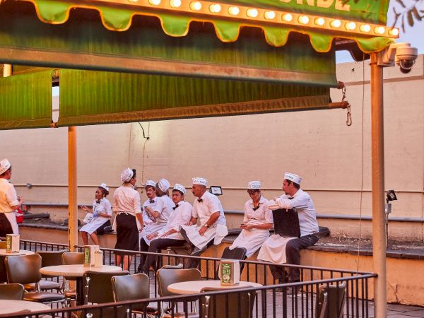 An outdoor seating area of Café Du Monde with several people in white uniforms sitting under an awning, and empty tables and chairs.