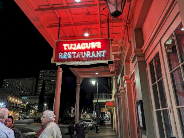 The image shows a street view with people and a lit sign reading "Tujague's Restaurant" under red neon lights at night.
