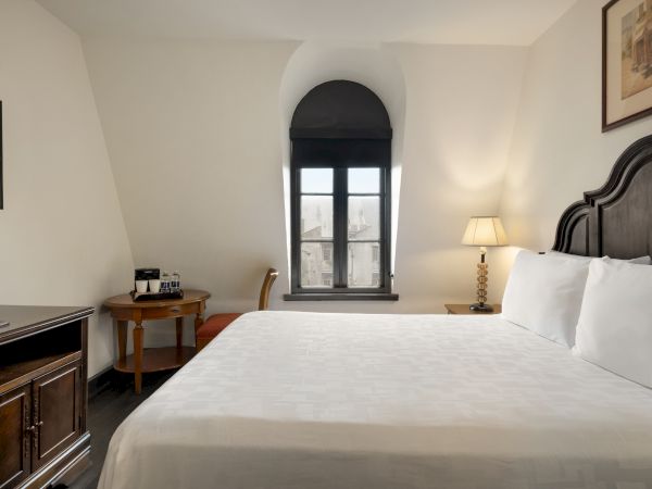 A cozy hotel room with a neatly made bed, bedside lamp, small desk, chair, and a window with a view; TV on the opposite wall.