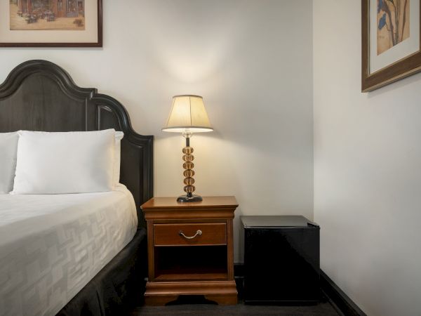 A cozy bedroom corner with a bed, nightstand, lamp, mini fridge, and wall art creates a warm and inviting atmosphere.