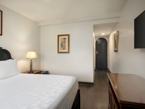 A hotel room with a bed, nightstand, lamp, paintings, TV, and wooden dresser; entrance visible in background.
