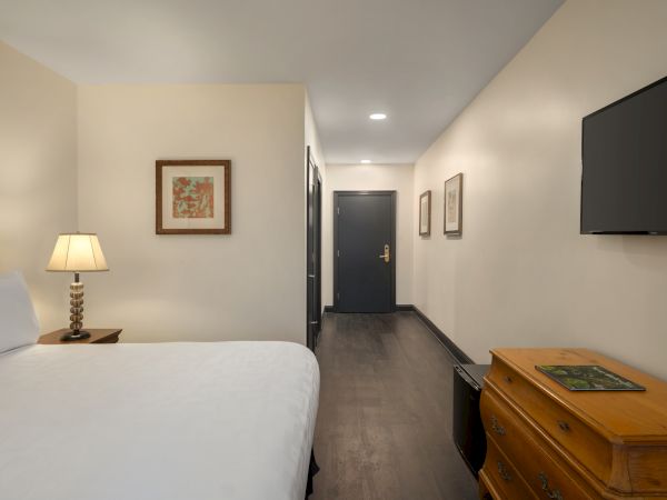 The room features a bed, a lamp, a wall-mounted TV, framed art, a wooden dresser, and a doorway leading out.