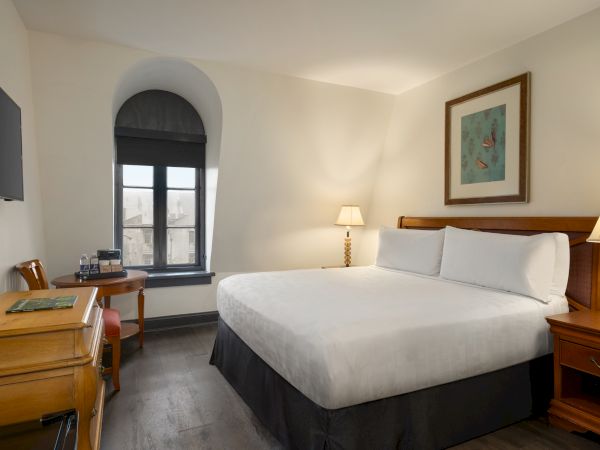 A hotel room with a bed, two lamps, a desk, chair, TV, and a window. The room has light-colored walls and a framed picture.