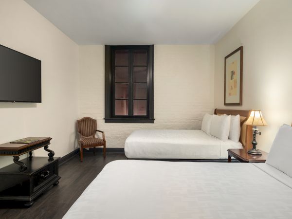 A hotel room with two beds, a wall-mounted TV, a wooden chair, a lamp on a nightstand, and a framed picture on the wall.