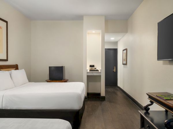 A hotel room with two beds, a TV, and a small shelf, featuring minimalist decor and wooden flooring.