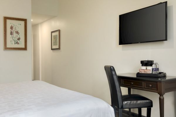 A tidy hotel room features a bed, wall-mounted TV, a desk with a chair, and artwork on light-colored walls.