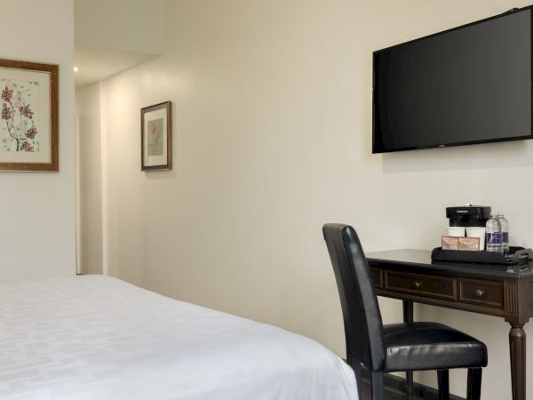 A tidy hotel room features a bed, wall-mounted TV, a desk with a chair, and artwork on light-colored walls.