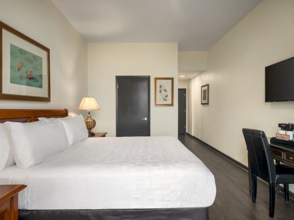 A hotel room with a large bed, wall art, a TV, desk with chair, lamp, and coffee maker on a cabinet. The decor is minimal and modern.