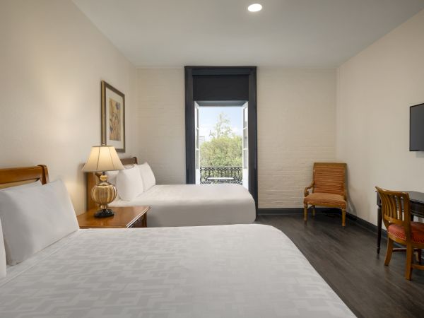 The image shows a hotel room with two beds, a lamp, a TV, a desk, and a chair. There's a window with a view outside.