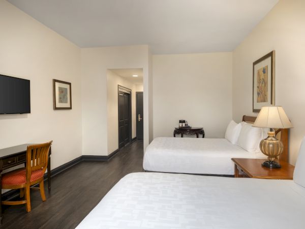 A hotel room with two beds, a small desk with chair, wall art, a lamp, and a TV. A hallway leads to a door.