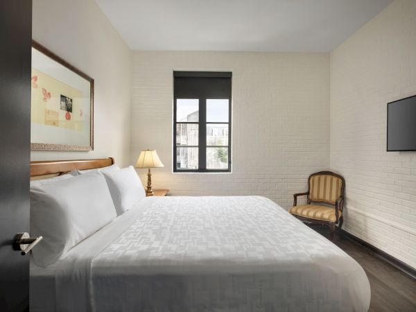 A cozy bedroom with a large bed, side table with a lamp, chair, framed artwork, window, and wall-mounted TV.