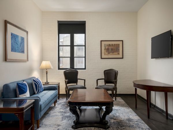 A cozy living room with a blue sofa, wooden coffee table, chairs, wall art, lamp, and TV, featuring a window with natural light.