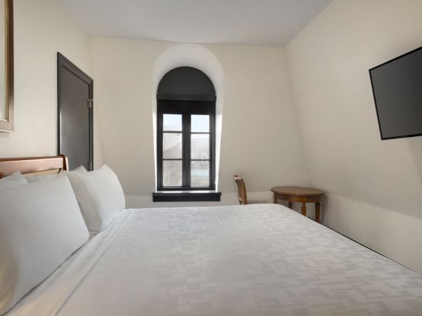 A minimalistic hotel room with a large bed, arched window, wall-mounted TV, and a small table and chair by the window.