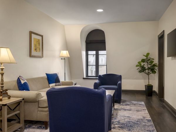 A cozy living room with blue chairs, a sofa, lamps, a plant, and a window; a serene space with soft lighting and decor.
