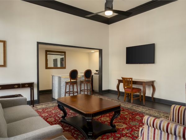The image shows a living room with a sofa, armchair, coffee table, TV, bar stools, and a desk, featuring a decorative rug and ceiling fan.