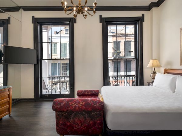 A stylish hotel room with a large bed, wooden dresser, TV, and windows overlooking a city balcony, featuring elegant decor.