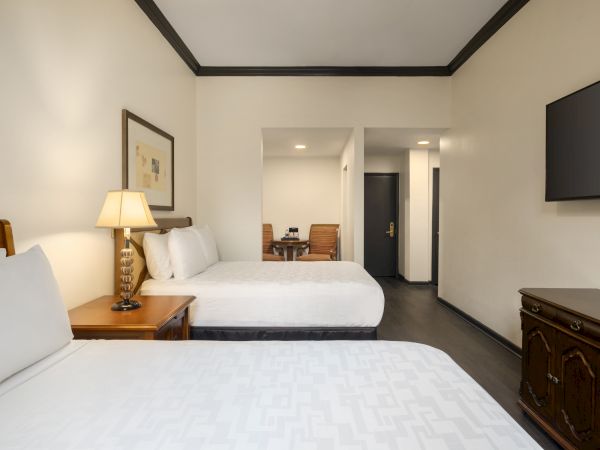 The image shows a hotel room with two beds, a lamp on a bedside table, a TV, and a small table with chairs in the background.